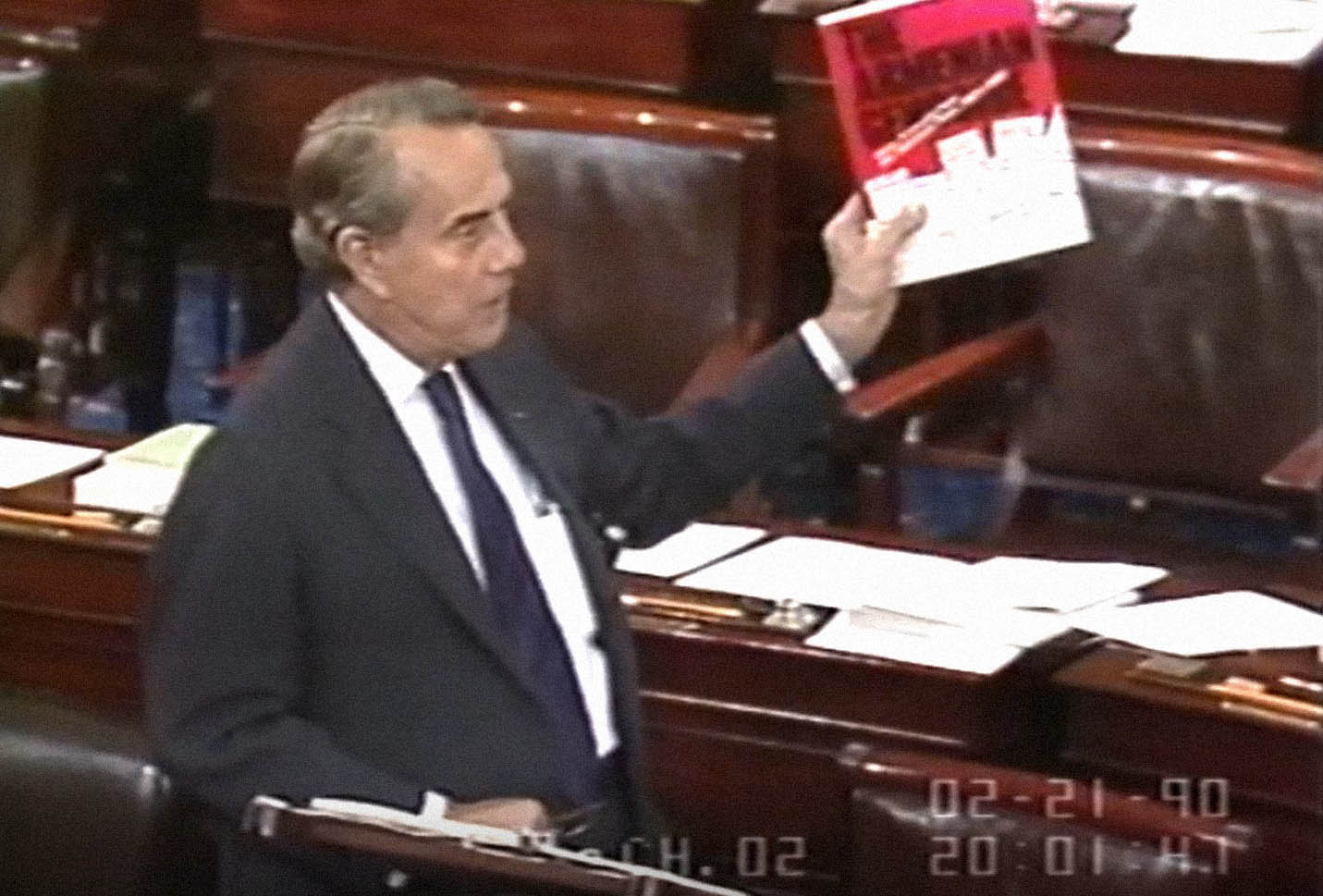 Senator Bob Dole stands on the Senate floor while talking and holding a book up that is titled 'The Armenian Genocide.'