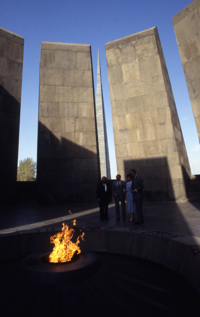 In the foreground, a large fire pit and flame sit within a round dropped section. In the background, four people stand in the shadow of a structure. Behind them, concrete pylons reach skyward, with gaps in between showing a blue sky.