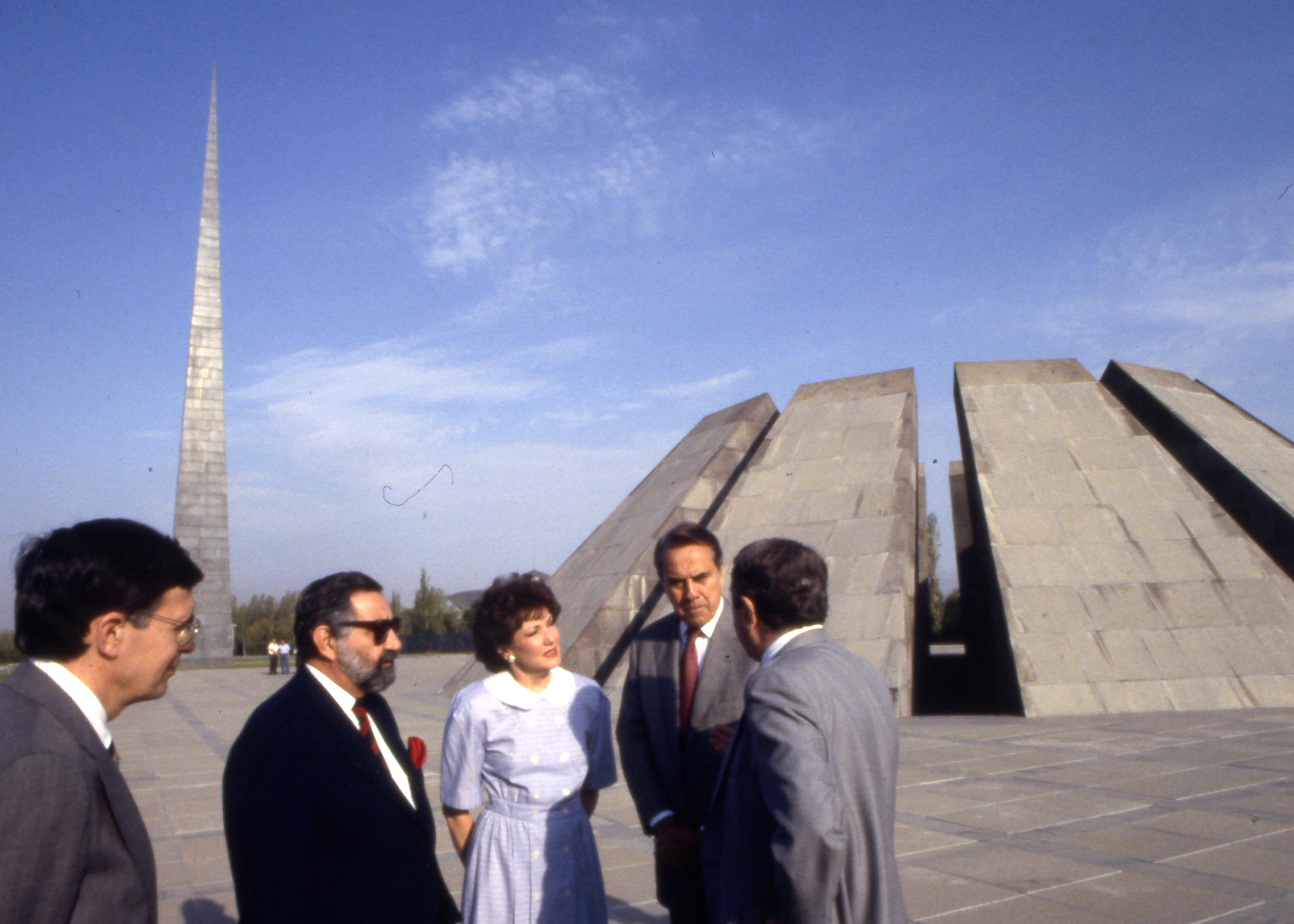 Three men and a woman stand talking near a large round concrete structure with deep slits around the sides, and another tall, pointed tower like structure.