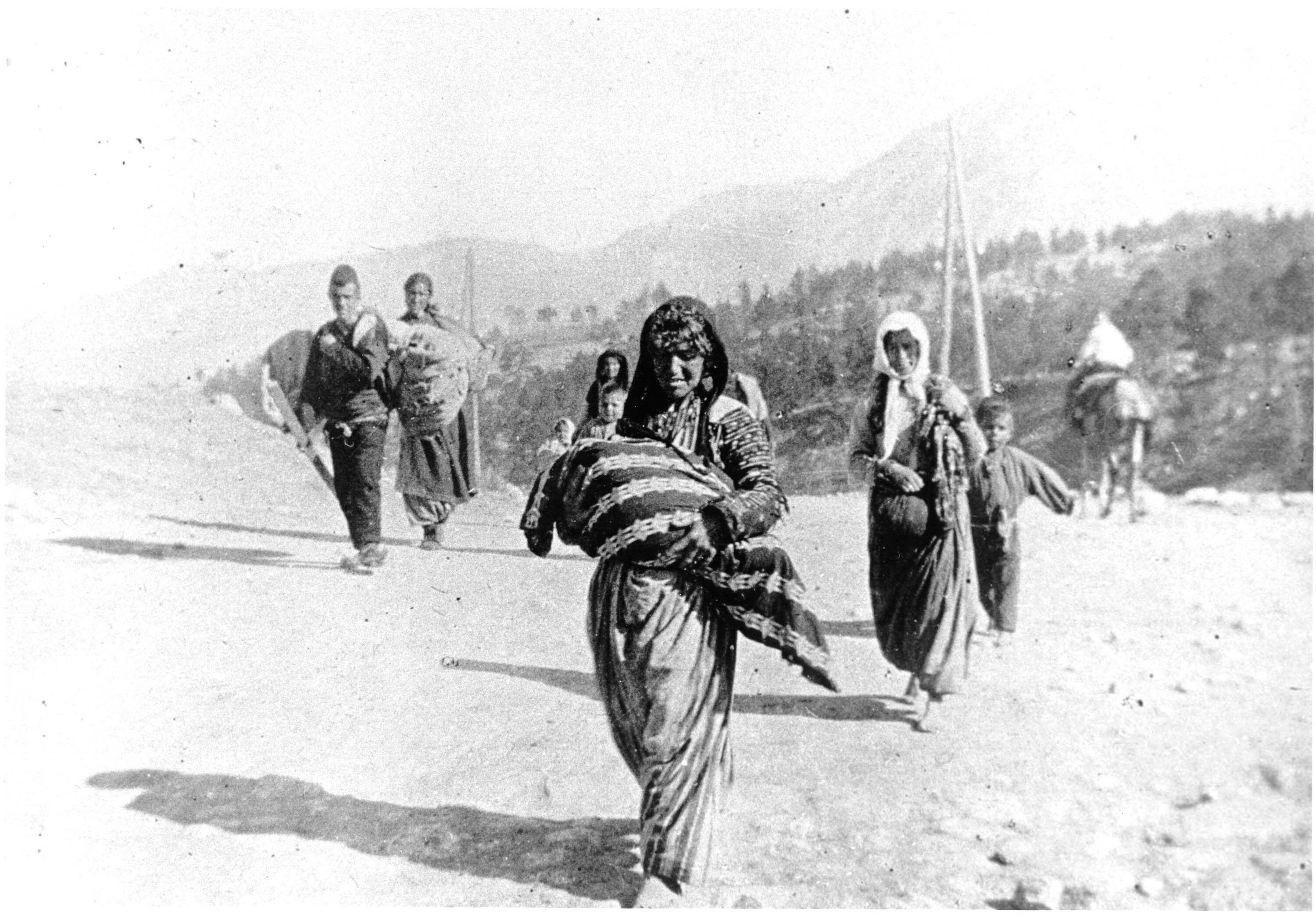 A group of men, women and children carrying bundles are walking toward the viewer, with mountainous forest in the background.